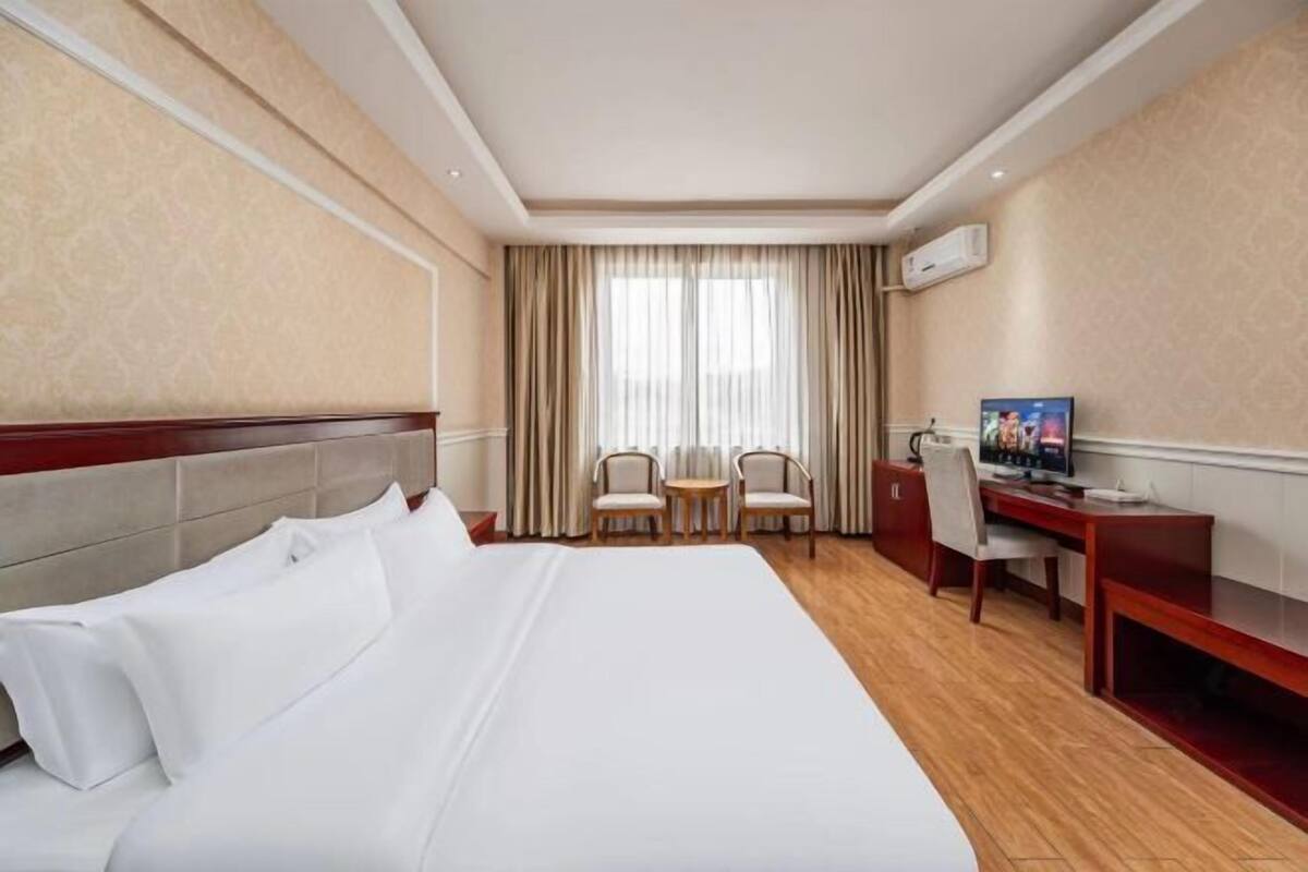 A hotel room with a large bed, a desk, two chairs, a TV, and a window with curtains.