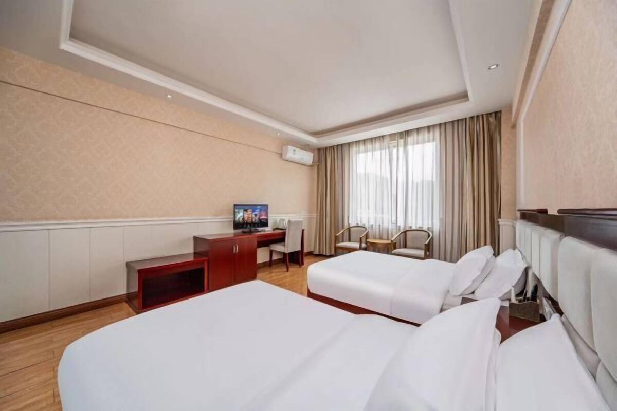 A hotel room with two beds, a desk, a TV, and a window with curtains.