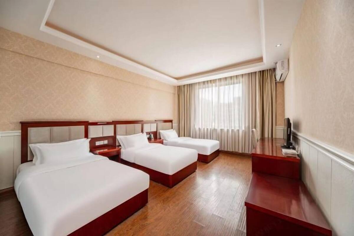 A hotel room with three beds, a wooden floor, a TV, and a window with curtains.