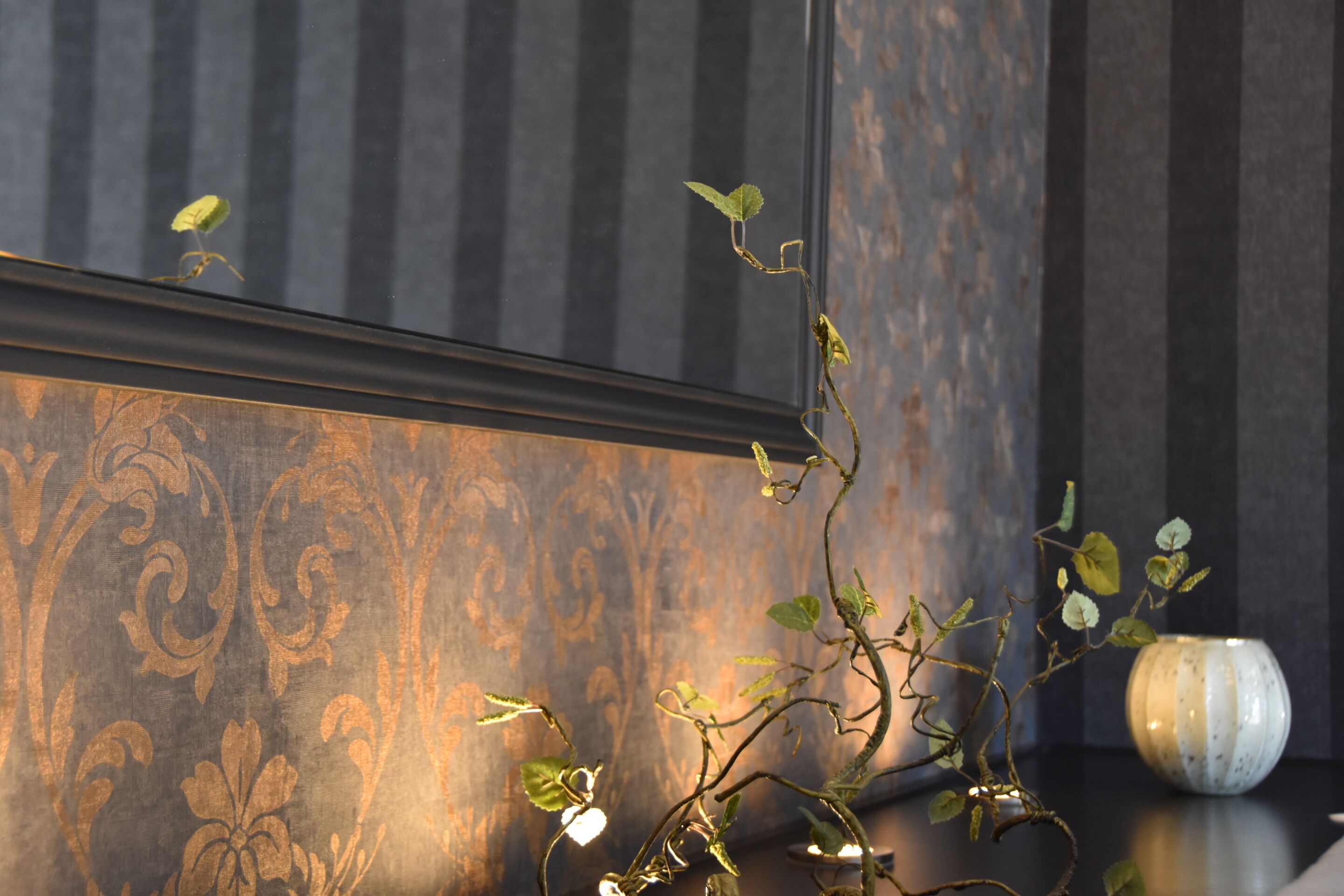 A dark wooden wall with a decorative pattern, a small vase, and a plant with green leaves and a twisted stem.