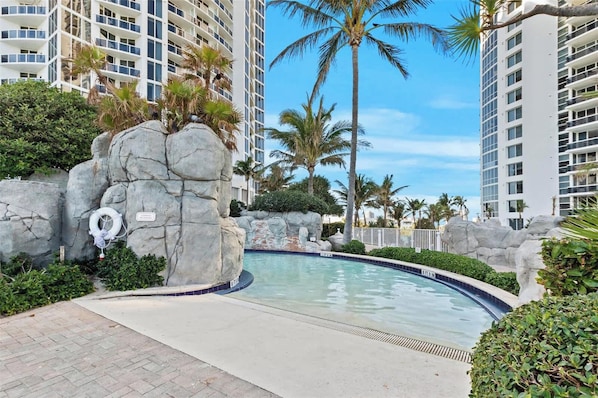 A swimming pool with a rock feature, surrounded by palm trees and a high-rise building in the background.