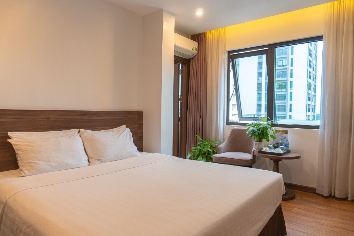 Una habitación de hotel con una cama grande, cabecera de madera, una mesita con una planta y una ventana con cortinas.