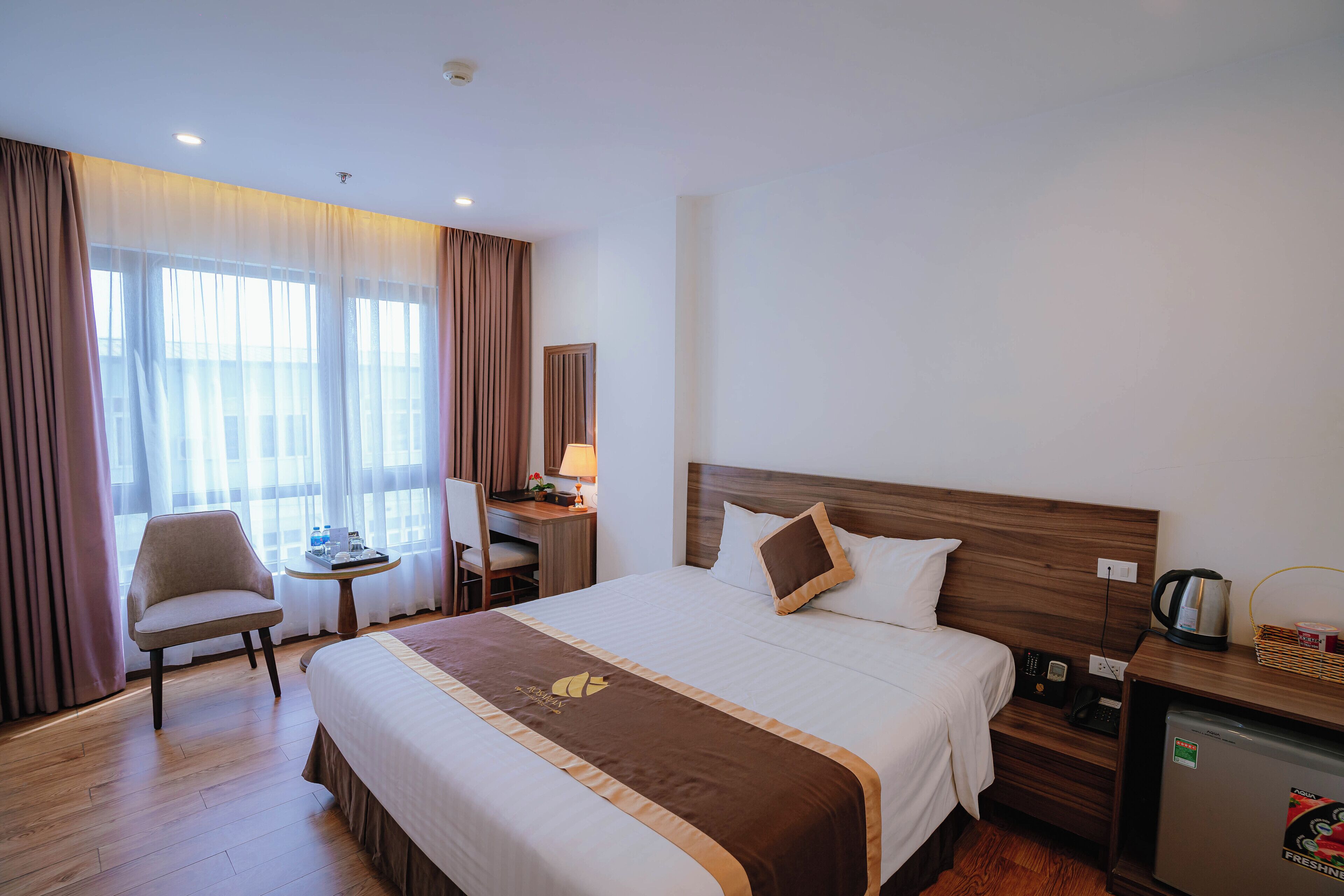 A hotel room with a large bed, a desk with a chair, a small table, and a refrigerator.