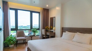 A hotel room with a large bed, a desk, a chair, and a view of the city through large windows. - A89 Rosarian Hotel (Hanoi)
