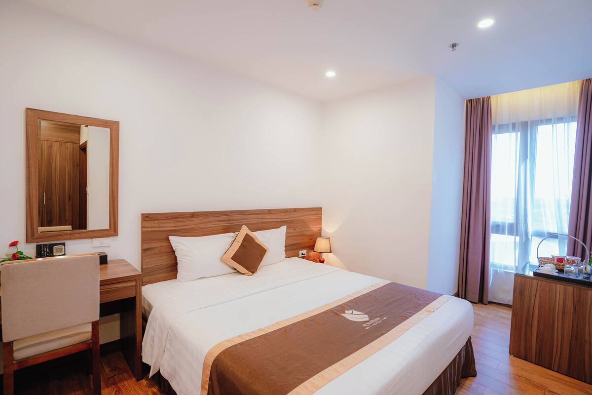 A hotel room with a large bed, a wooden desk, a chair, a nightstand with a lamp, a mirror, and a window with curtains.