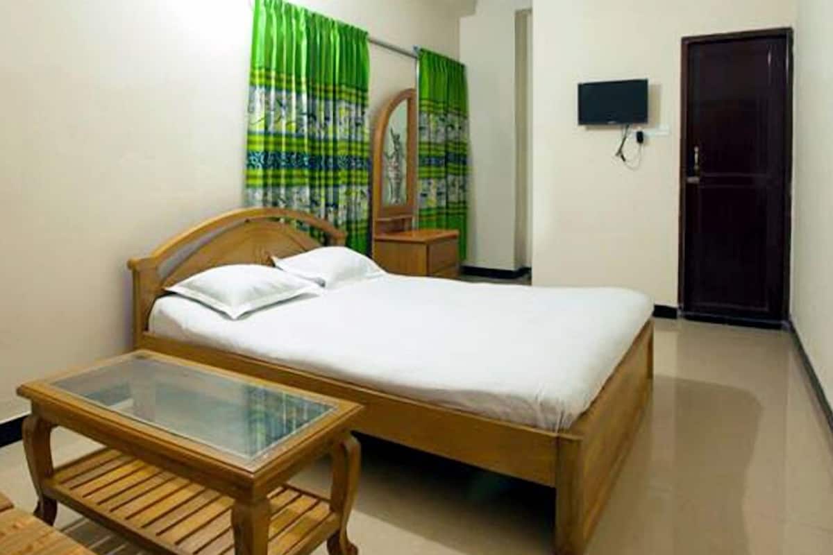 A single bed with a wooden frame and white bedding, a small wooden table with a glass top, a mirror on a wooden stand, and a television mounted on the wall.
