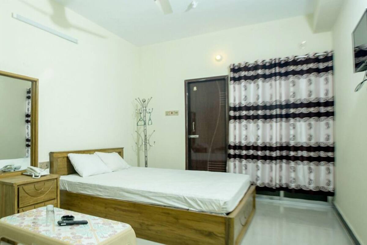A bedroom with a bed, a bedside table with a phone and a remote, a mirror, a wardrobe, and a curtain.