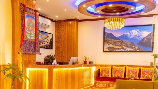 Um saguão de hotel com balcão de recepção em madeira, um sofá, um candelabro e uma pintura com paisagem de montanhas na parede.