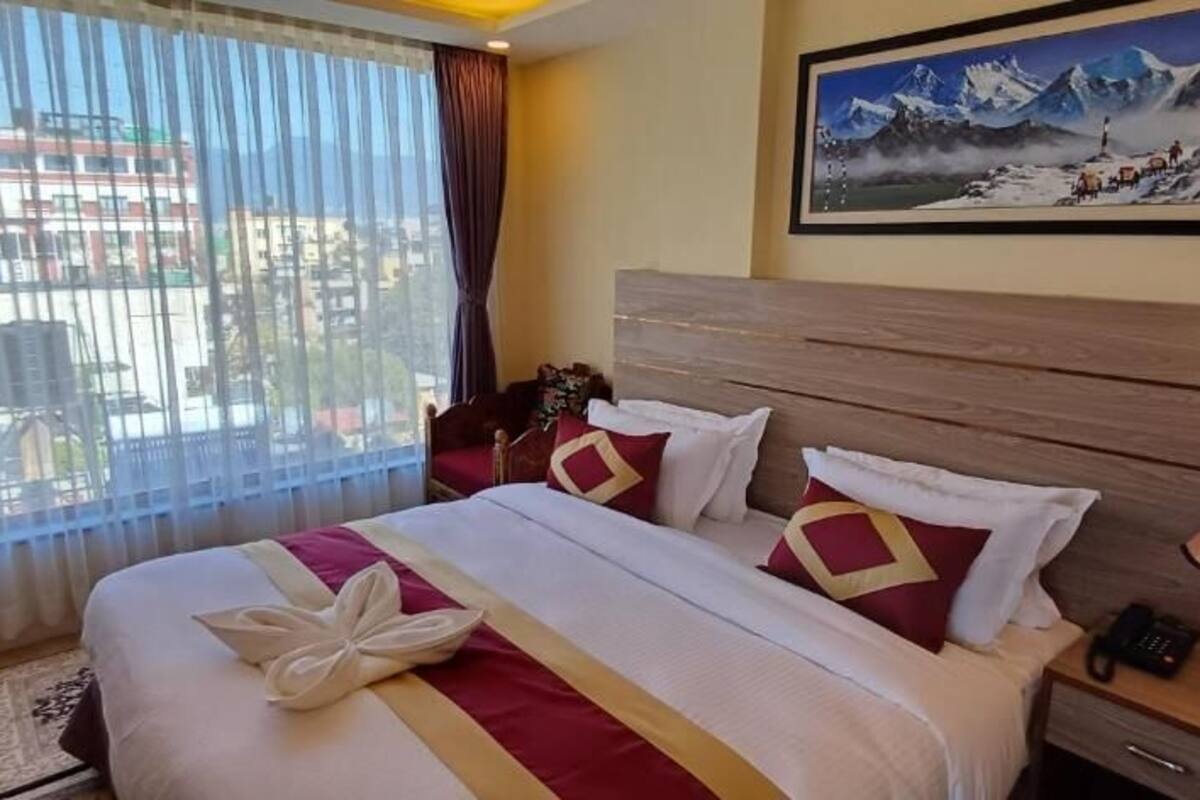 A hotel room with a large bed, a nightstand with a lamp, a painting on the wall, and a view of buildings through the window.