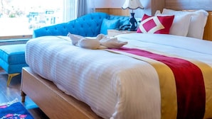 Quarto de hotel com uma cama grande, uma poltrona azul, uma cabeceira de madeira, uma janela com cortinas e um quadro na parede.