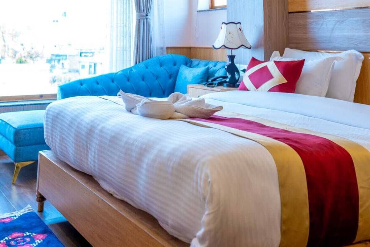A hotel room with a large bed, a blue armchair, a wooden headboard, a window with curtains, and a painting on the wall.