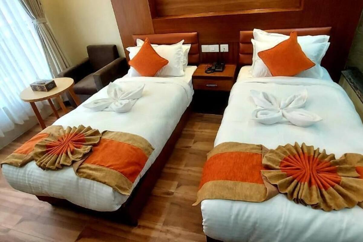 A hotel room with two beds, each with a white comforter, orange and brown decorative pillows, and a neatly folded white towel. There is a wooden headboard, a small round table, and a chair.