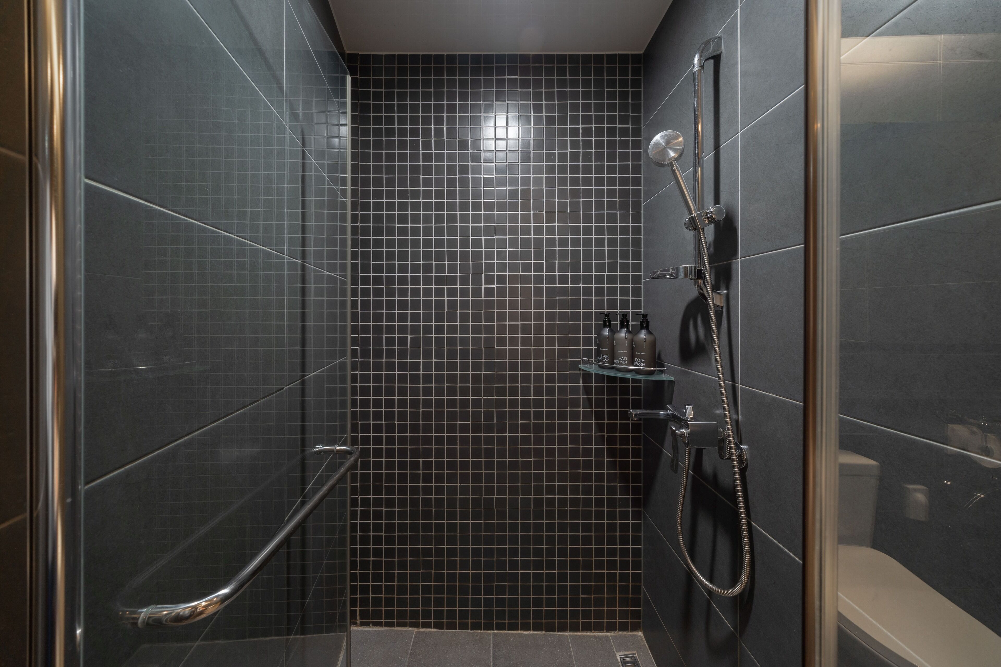 A modern bathroom with dark tiled walls, a glass shower enclosure, a handheld showerhead, and a small shelf with toiletries.