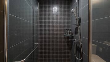 A modern bathroom with dark tiled walls, a glass shower enclosure, a handheld showerhead, and a small shelf with toiletries.