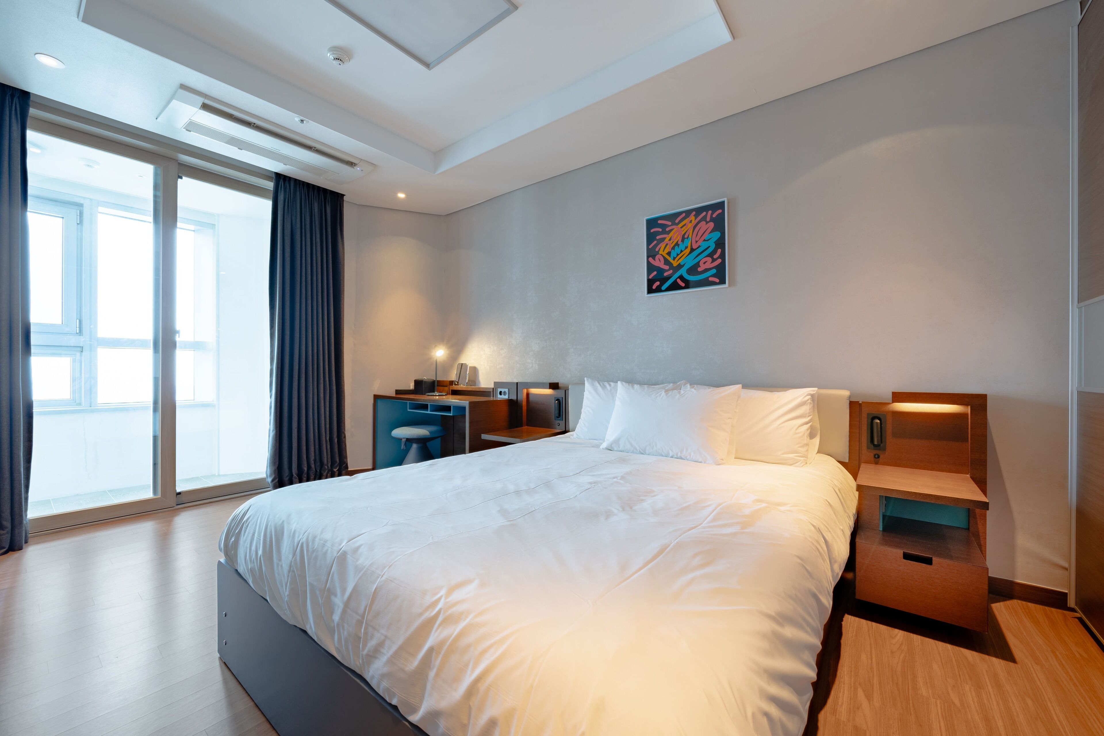 Una habitación de hotel moderna con una cama grande, un escritorio, una silla, una mesita de noche y un ventanal con cortinas.