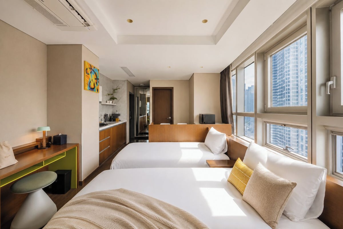 Habitación de hotel con dos camas, un escritorio, una silla, una ventana con vista a la ciudad y una puerta que da a un pasillo.