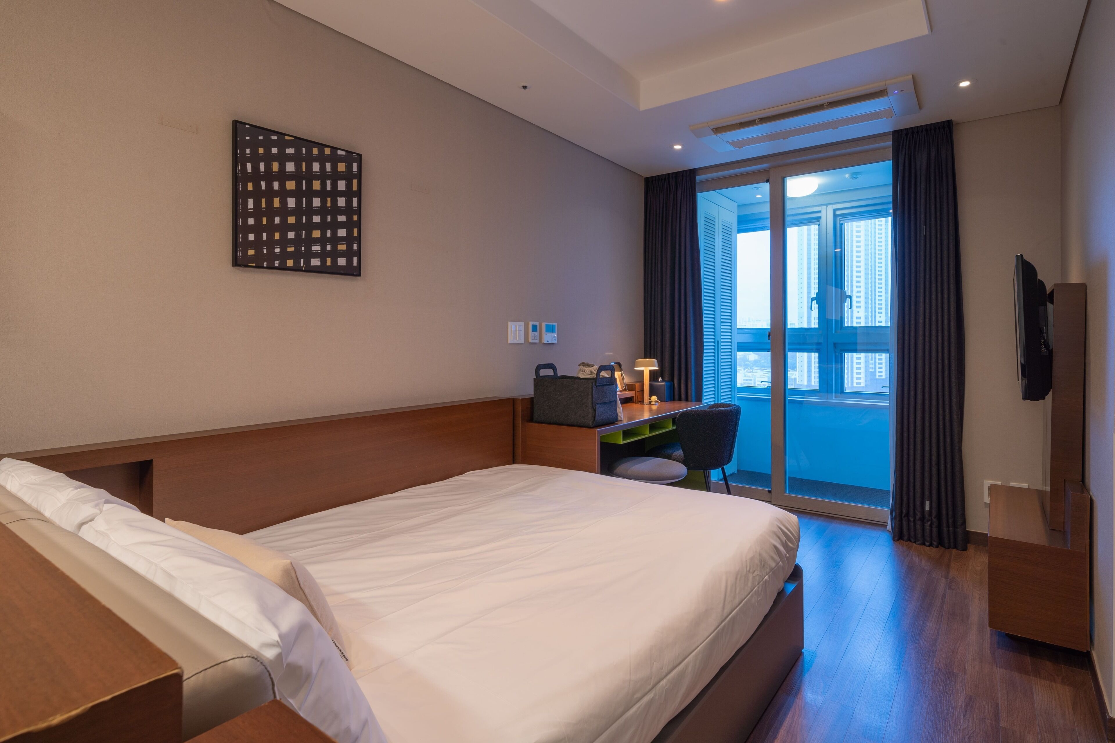 A hotel room with a large bed, a desk with a chair, a TV, and a view of the city through the window.