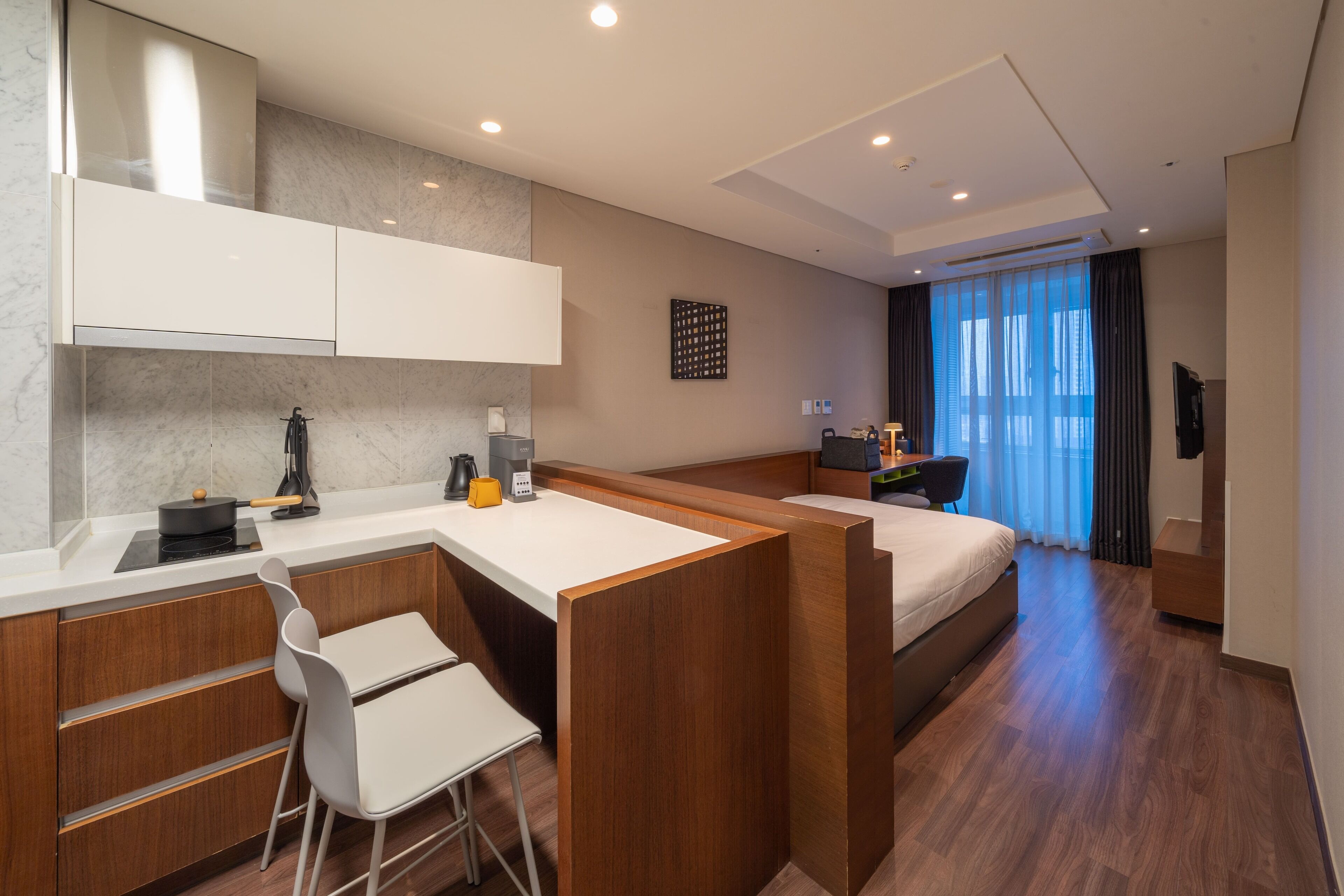 A modern hotel room with a kitchenette, a bed, a desk, and a large window with curtains.