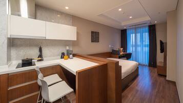 A modern hotel room with a kitchenette, a bed, a desk, and a large window with curtains.