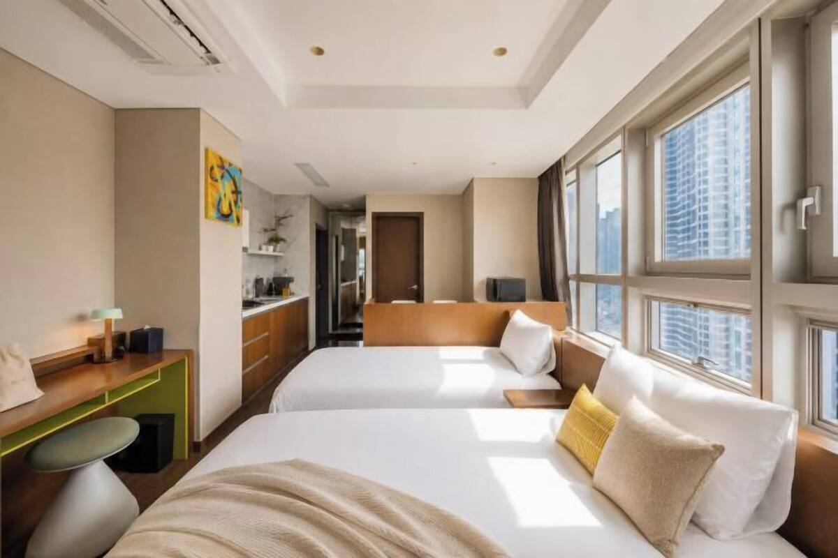 Habitación de hotel con dos camas, un escritorio, una silla y ventanas amplias.