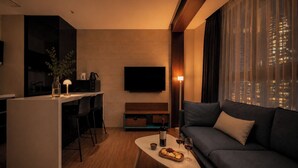 Una sala de estar moderna con un sofá, una mesa de centro con comida y bebidas, un televisor y una zona de cocina con una planta y una lámpara.