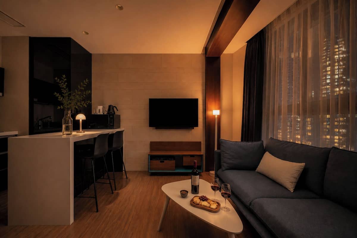Una sala de estar moderna con un sofá, una mesa de centro con comida y bebidas, un televisor y una zona de cocina con una planta y una lámpara.