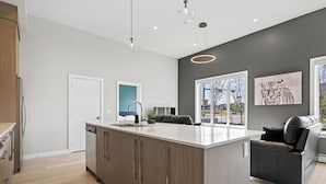 A modern kitchen with a central island, wooden cabinets, and a black leather sofa in a living area with large windows. - Ground-Level 3BR Condo w/ UG Parking, Lawn Access (Northeast)