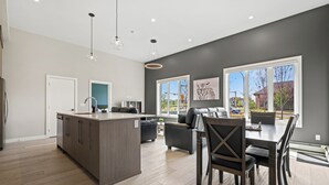 A modern kitchen with a central island, a dining area with a dark wood table and chairs, and a living area with a sofa and coffee table. - Ground-Level 3BR Condo w/ UG Parking, Lawn Access (Northeast)