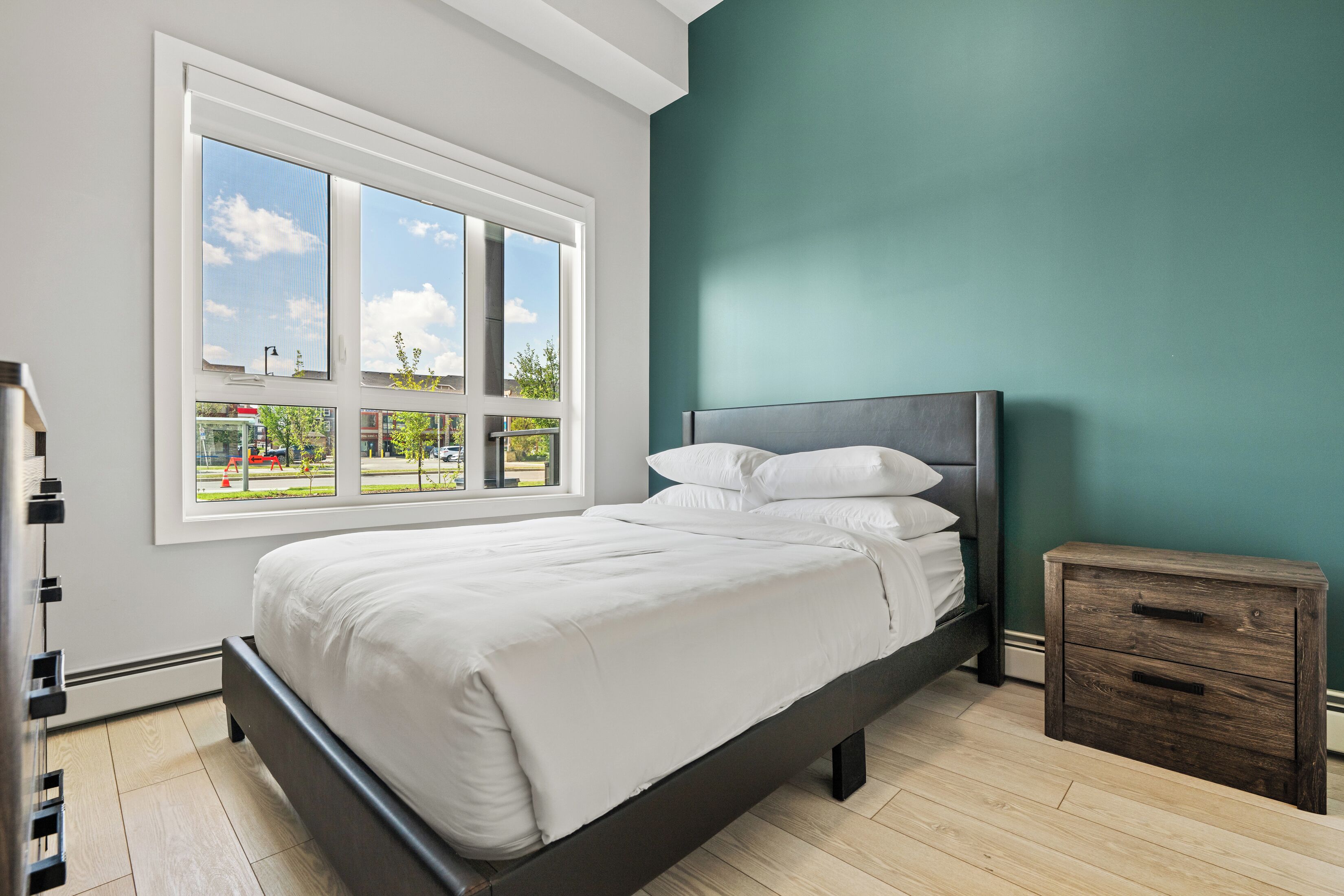 Une chambre avec une grande fenêtre, un lit, une table de chevet et un mur de couleur turquoise.