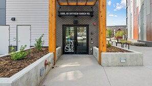 A building entrance with a wooden pergola, a sign reading "1000, 60 SKYVIEW RANCH RD.", and a glass door with a logo. - Ground-Level 3BR Condo w/ UG Parking, Lawn Access (Northeast)
