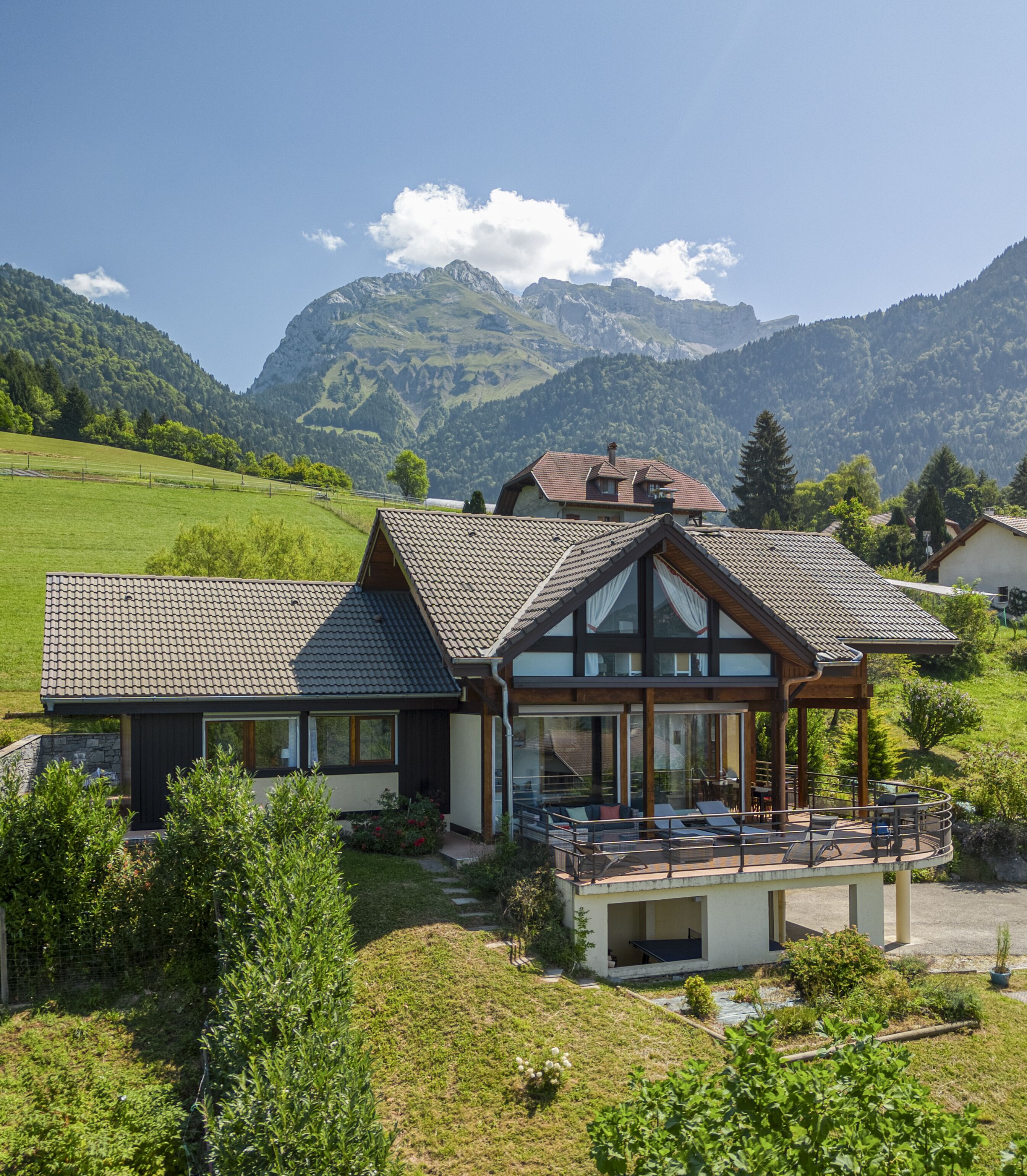 A modern house with a large balcony, surrounded by lush greenery and mountains in the background.