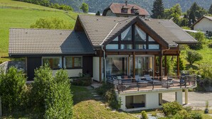 A modern house with a large balcony, surrounded by lush greenery and mountains in the background.