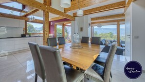 A spacious dining area with a wooden table and chairs, large windows offering mountain views, and a kitchen area with white cabinets.