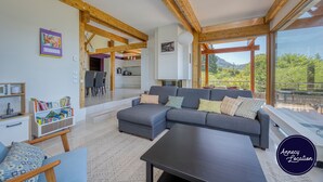 A modern living room with a grey sectional sofa, wooden beams, a dining area, and a view of greenery.