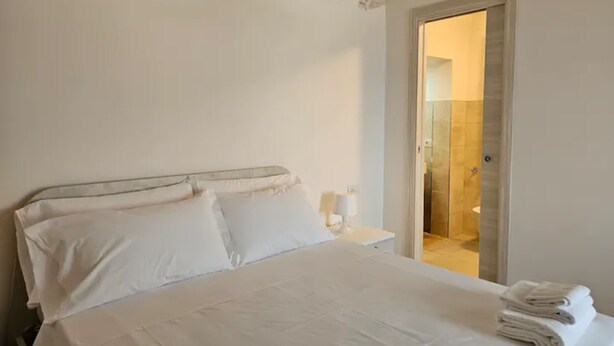 Habitación de hotel con una cama grande, ropa de cama blanca, una pared decorativa de piedra y un baño visible a través de una puerta abierta.