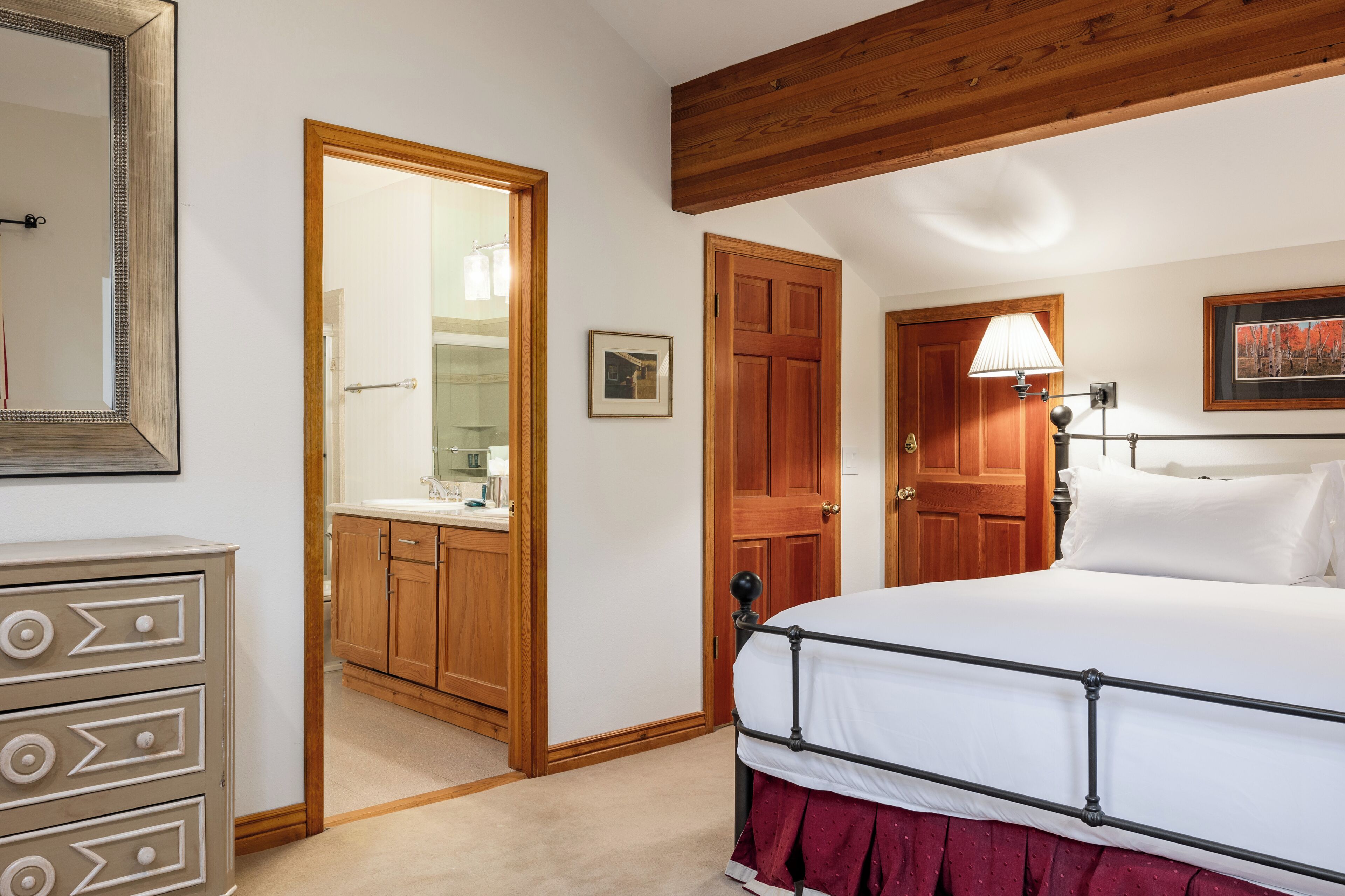 Un dormitorio con techo de madera, cama con ropa de cama blanca, puerta de madera y baño visible a través de una puerta abierta.