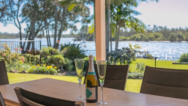 A table with a bottle of wine and two glasses, set outdoors with a view of a lake and palm trees.