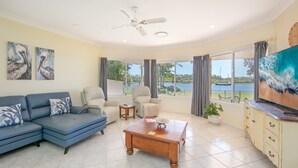A living room with a blue sofa, two armchairs, a wooden coffee table, and a TV on a cabinet. There are paintings on the wall and a view of a lake with boats.