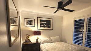 Ein Schlafzimmer mit einem Bett, einem Deckenventilator, einem Nachttisch mit einer Statue und einem Fenster mit Jalousien.