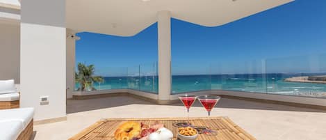 A balcony with a dining table set with food and drinks, overlooking a beach and ocean.