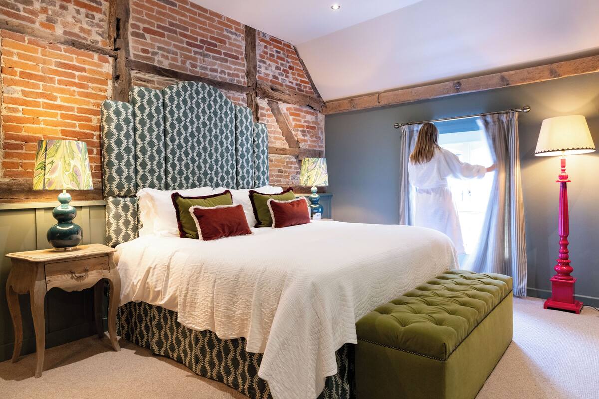 A bedroom with a large bed, a green ottoman, a nightstand with a lamp, and a brick wall.