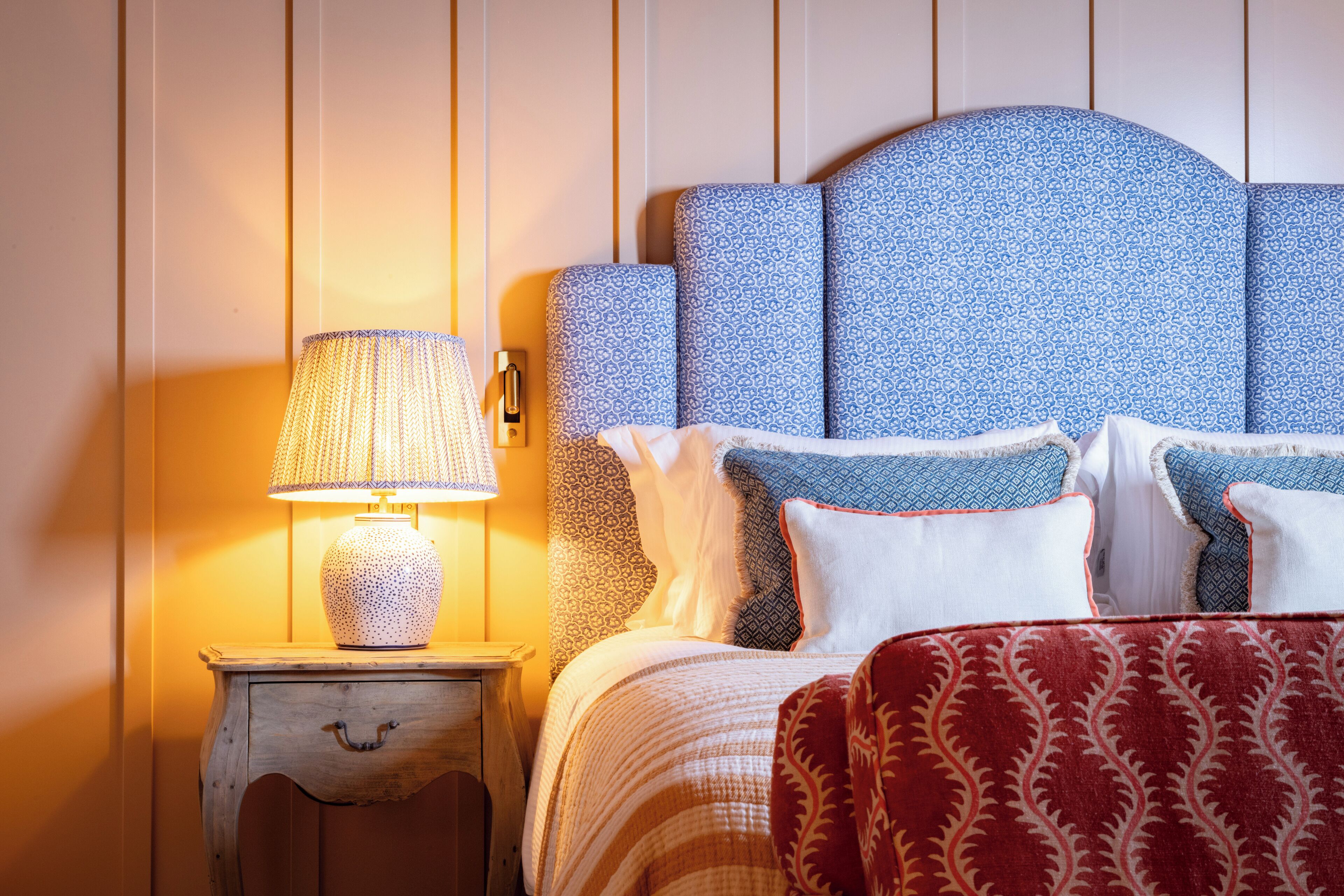 Een net opgemaakt bed met gestreepte kussens en een rood-witte deken, een bedlampje en een houten nachtkastje met een lade.