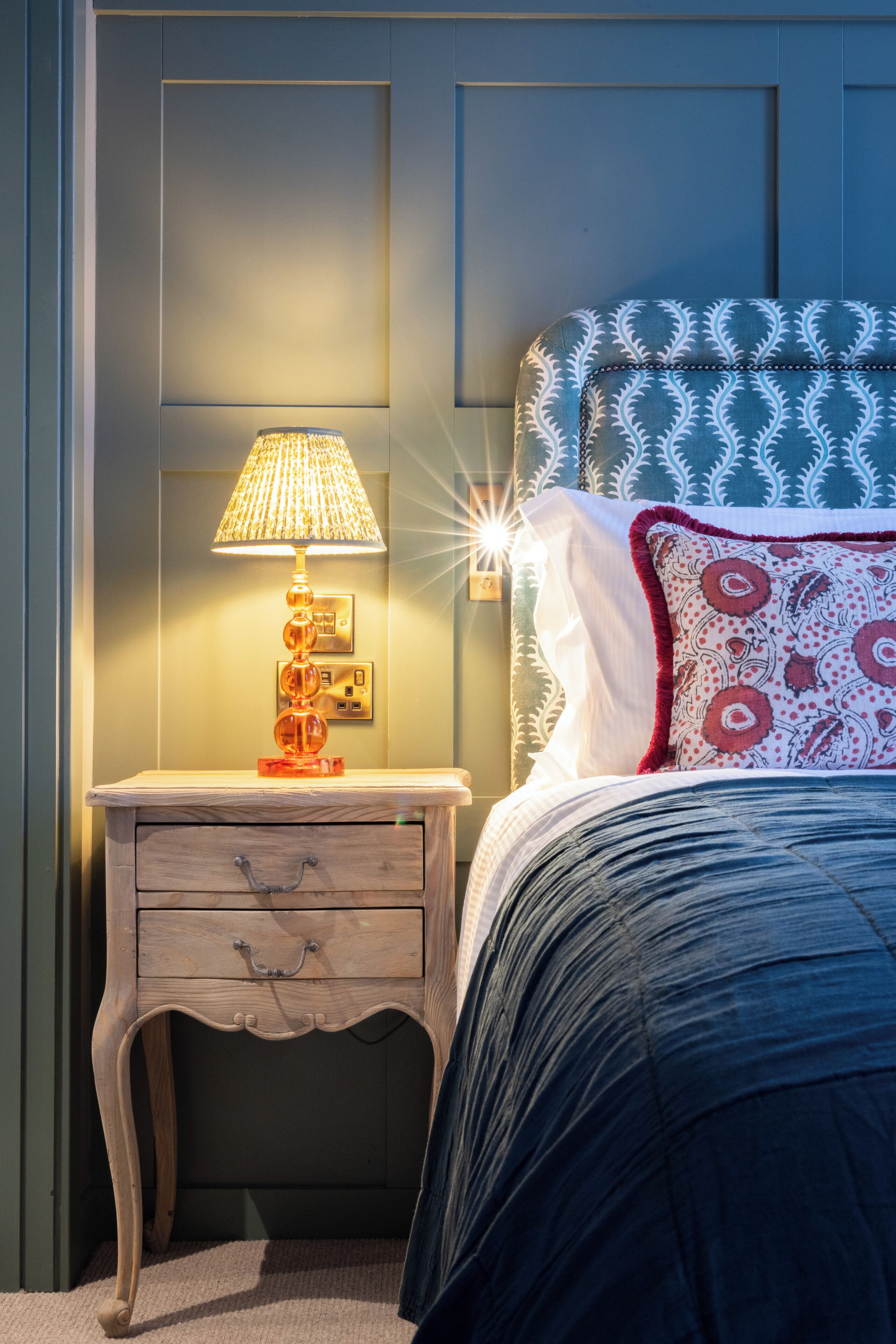 A neatly made bed with a patterned headboard, a bedside lamp, and a wooden nightstand with drawers.