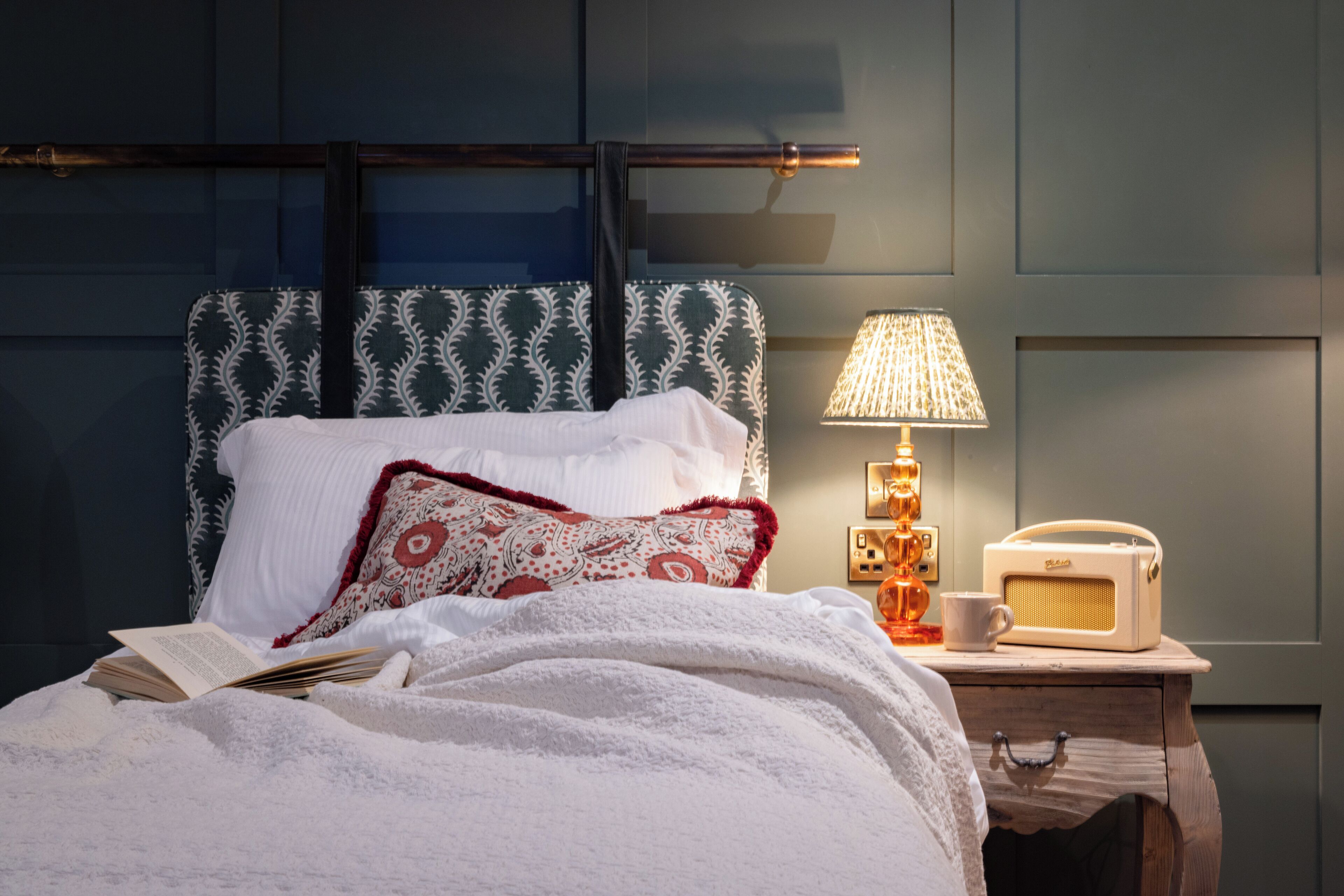 Een net opgemaakt bed met witte lakens en een hoofdbord met patroon, een bedlamp, een radio en een kopje op een houten nachtkastje.