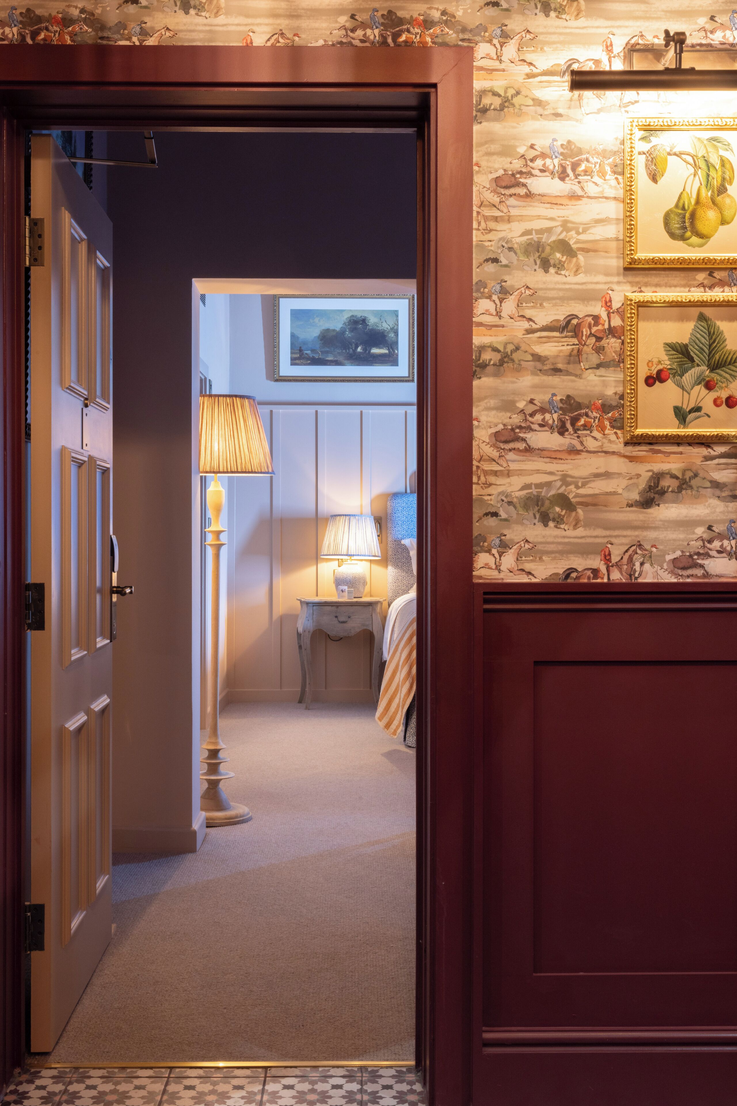 A hallway with a wooden door, a lamp, a small table with a lamp, and a wall with framed pictures.