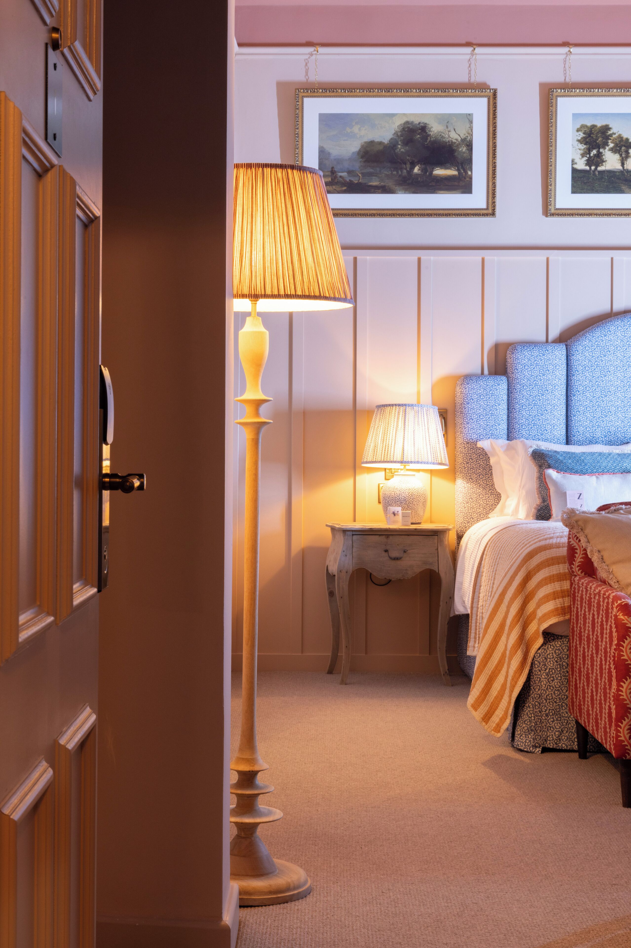 Habitación de hotel con puerta de madera, una lámpara de pie, una mesita de noche con lámpara, una cama con ropa de cama estampada y dos cuadros en la pared.