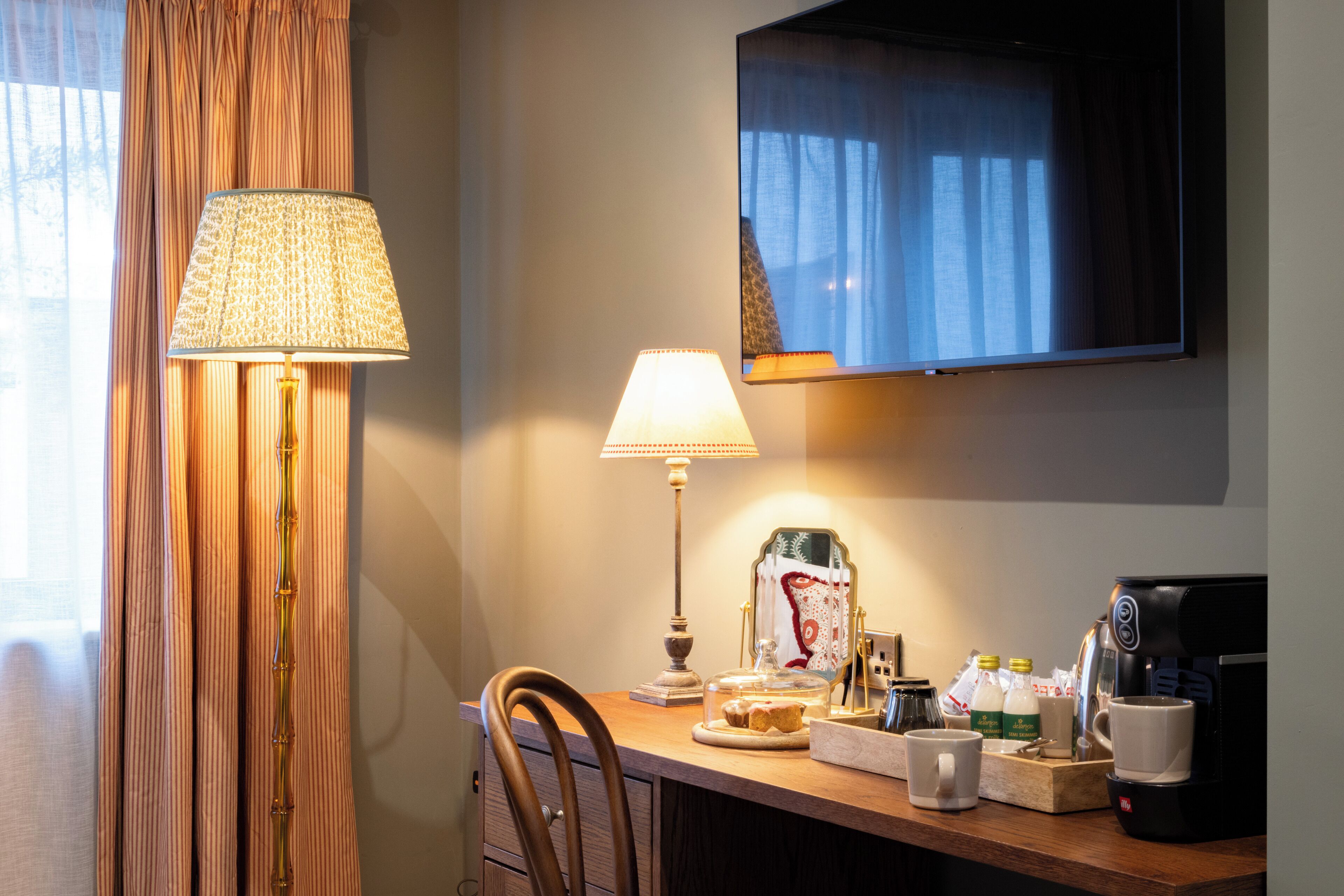 A hotel room with a desk, a lamp, a coffee maker, and a television.