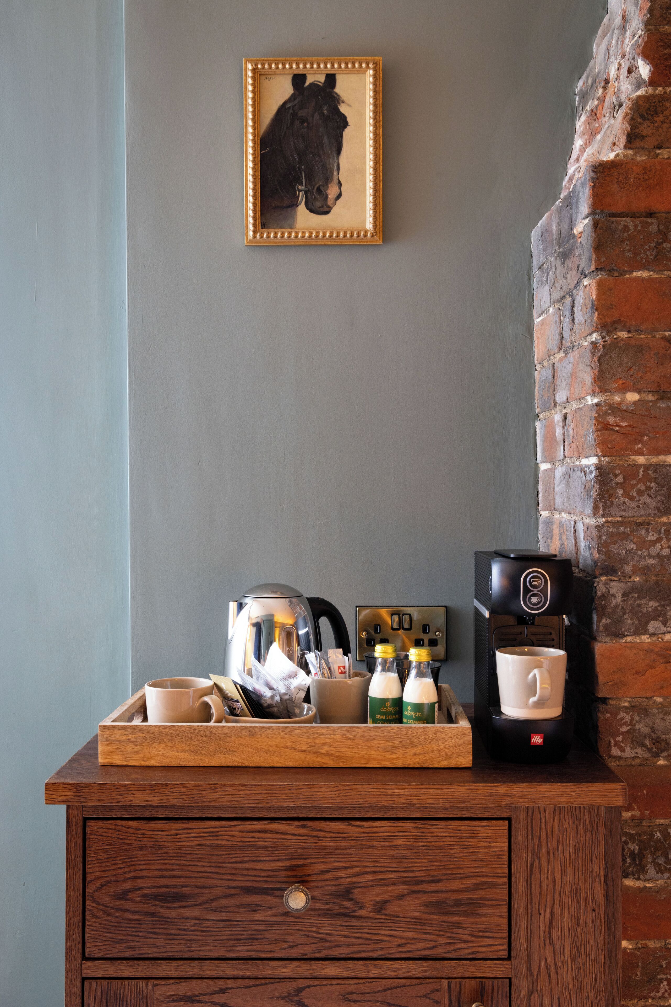 A wooden tray with coffee cups, a coffee maker, and a framed picture on a wall.