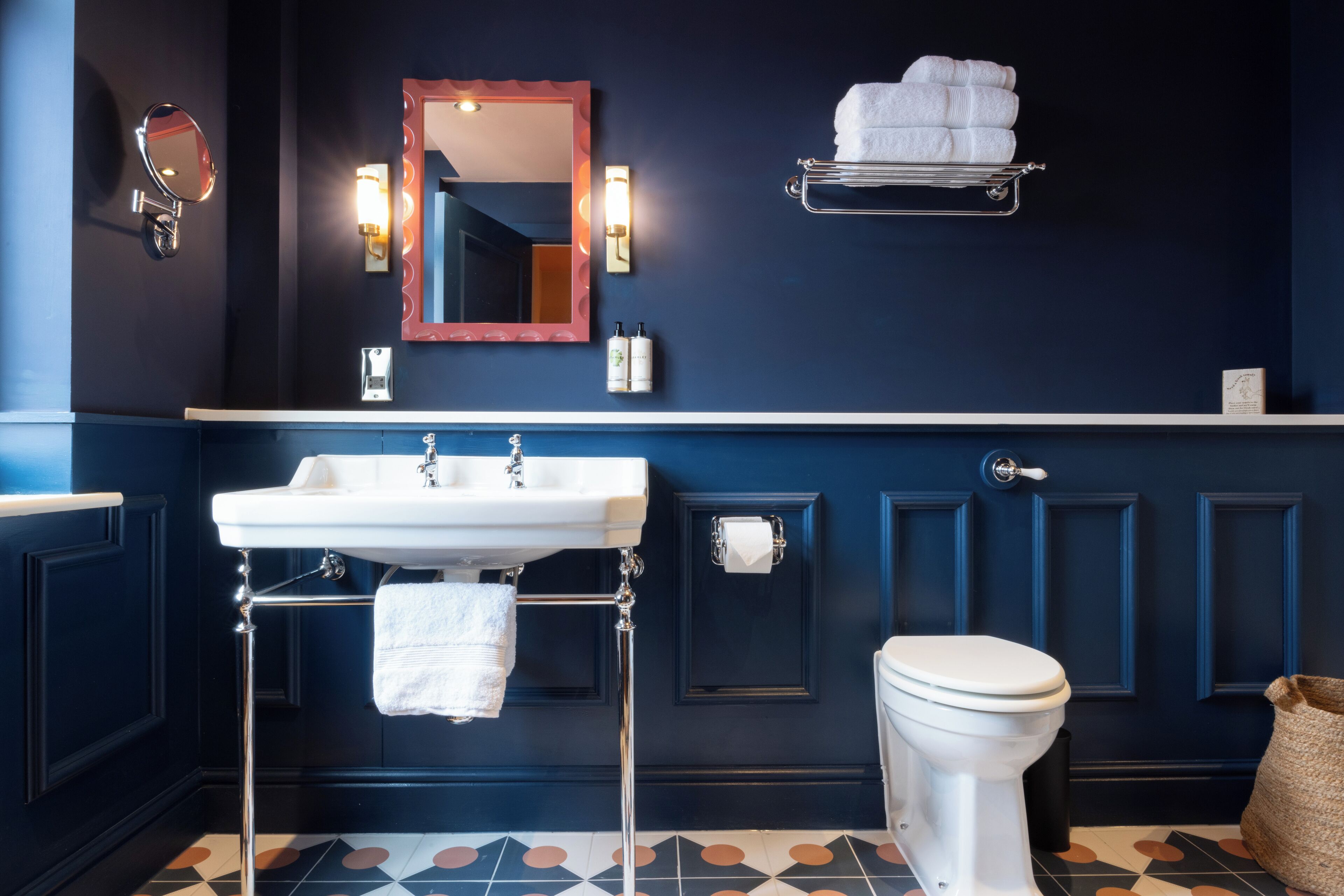 Ein Badezimmer mit einem weißen Waschbecken, Spiegel und Toilette. Es gibt einen Handtuchhalter mit Handtüchern, eine Seifenspender und ein Toilettenpapierhalter. Der Boden ist mit einem geometrischen Fliesenspiegel versehen.