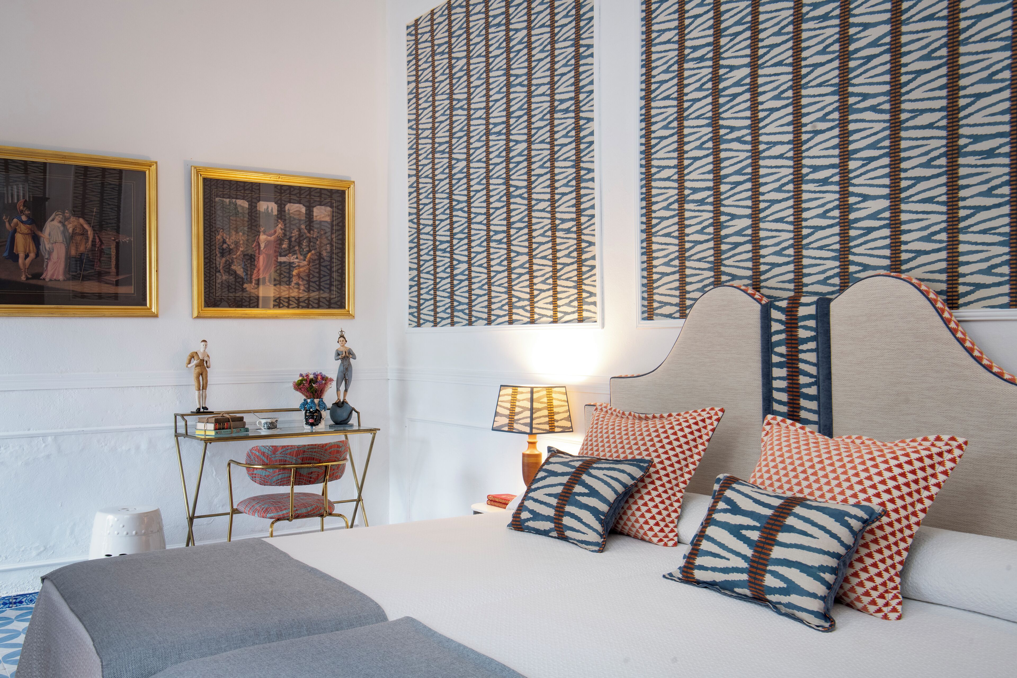 Un dormitorio ordenado con cama, almohadas, una mesita de noche, lámpara y dos cuadros enmarcados en la pared.